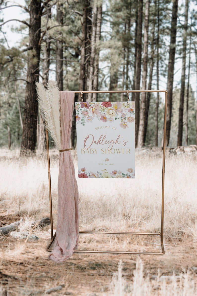 May include: A gold metal stand with a sign that says "Welcome to Oakleigh's Baby Shower April 22, 2023". The sign has a floral design with pink, yellow, and green flowers. The stand is in front of a forest background.