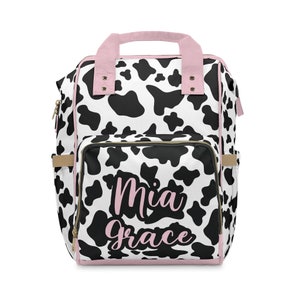 May include: A black and white cow print nappy bag with pink accents and the name "Mia Grace" written in pink script.