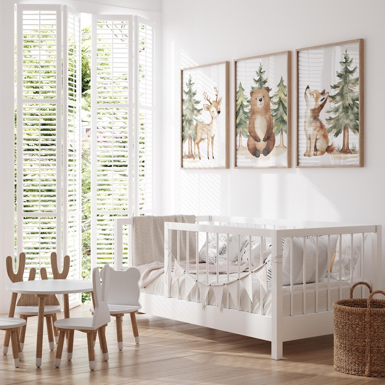 Op de afbeelding: Een lichte kinderkamer met een witte wieg, een kleine tafel en stoelen en drie ingelijste bosdierprints. De prints tonen een hert, een beer en een vos in een bosrijke omgeving. Een rieten mand staat op de vloer.