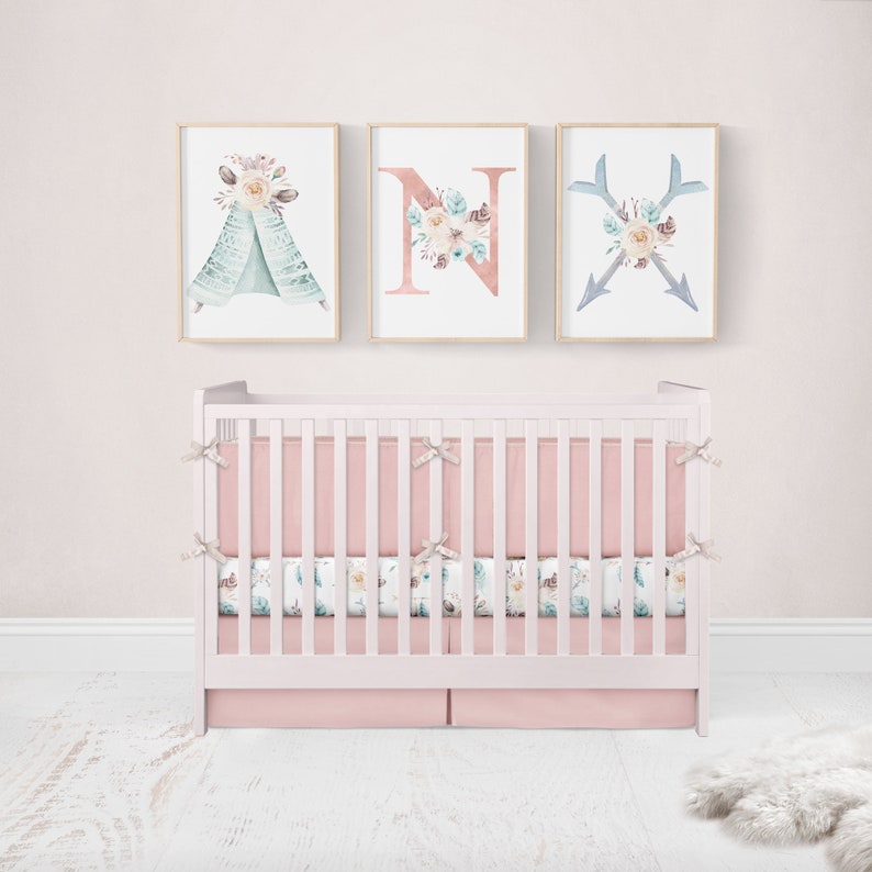 May include: A pink and white crib with a floral print crib skirt and matching bedding. Three framed prints with watercolor floral designs hang above the crib. The prints feature a teepee, the letter N, and two arrows.