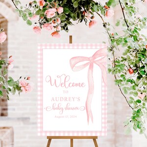May include: Pink and white gingham welcome sign with a pink bow and the text "Welcome to Audrey's Baby Shower August 17, 2024".