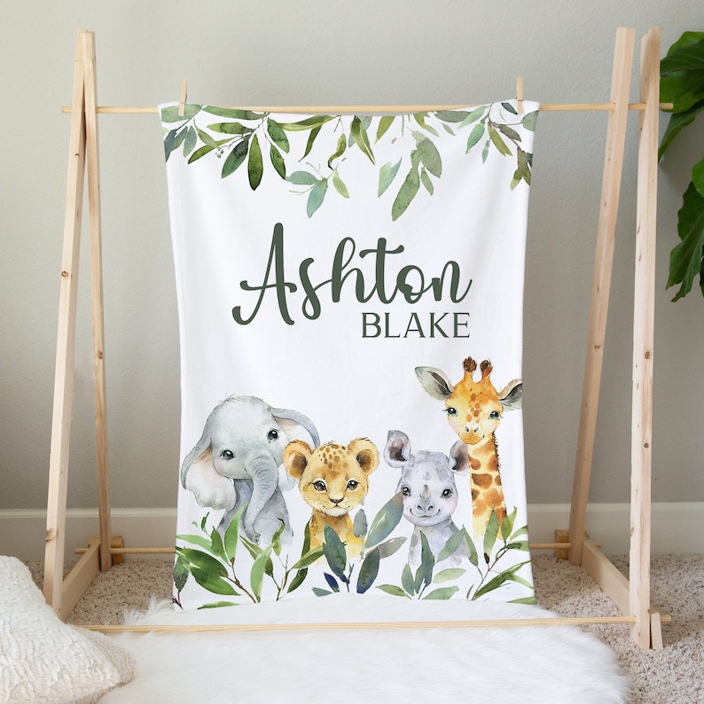 May include: A white baby blanket with a green leafy border and a watercolor illustration of a giraffe, elephant, lion, and rhinoceros. The blanket is personalized with the name "Ashton Blake".