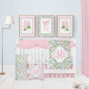 Puede incluir: Una cuna blanca con falda de volantes rosa y una colcha con estampado floral que lleva el monograma GMB. El conjunto incluye un coj&iacute;n cuadrado a juego con el monograma GMB, un panel lateral floral y tres cuadros de pared con hortensias y un lazo.