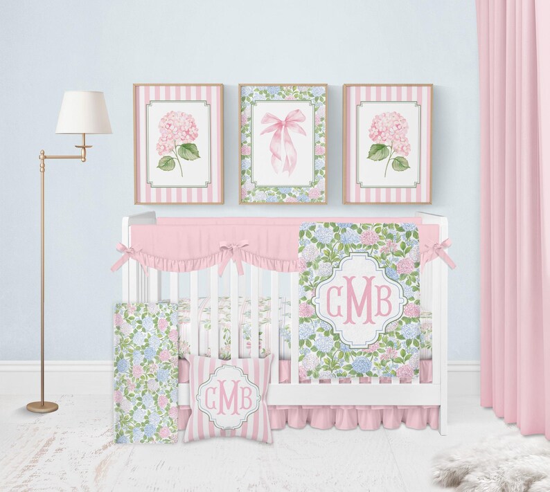 Puede incluir: Una cuna blanca con falda de volantes rosa y una colcha con estampado floral que lleva el monograma GMB. El conjunto incluye un coj&iacute;n cuadrado a juego con el monograma GMB, un panel lateral floral y tres cuadros de pared con hortensias y un lazo.