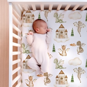 May include: A white cot sheet with a whimsical dragon and castle design. The sheet features watercolour illustrations of green and brown dragons, castles, and clouds on a white background.