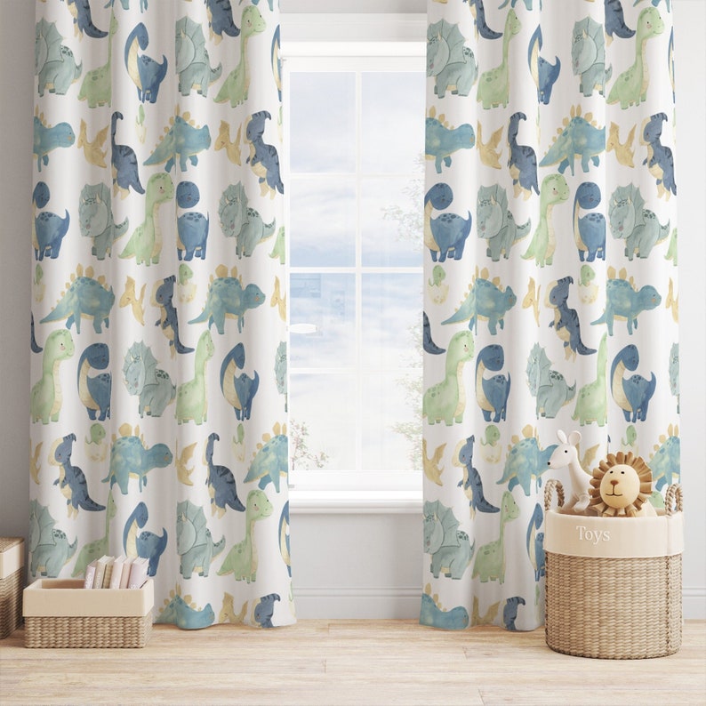 May include: A pair of white curtains with a repeating pattern of blue and green dinosaurs. The curtains are hung on a window with a view of a blurry outdoor scene. A wicker basket labeled "Toys" sits on the floor in front of the curtains, containing a stuffed lion and a stuffed rabbit.