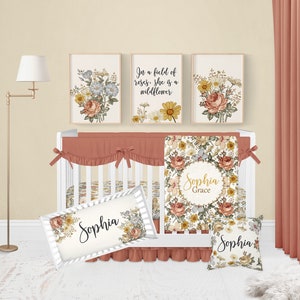Op de afbeelding: Een wit babybedje met een bloemenbeddengoedset. De beddengoedset bevat een deken, een kussen en een bumper. Het babybedje is versierd met drie ingelijste bloemenprints. De prints tonen een bloemenpatroon met de tekst "In een veld vol rozen is ze een wilde bloem". Het babybedje is ook versierd met een kussen met de tekst "Sophia Grace" en een kussen met de tekst "Sophia".