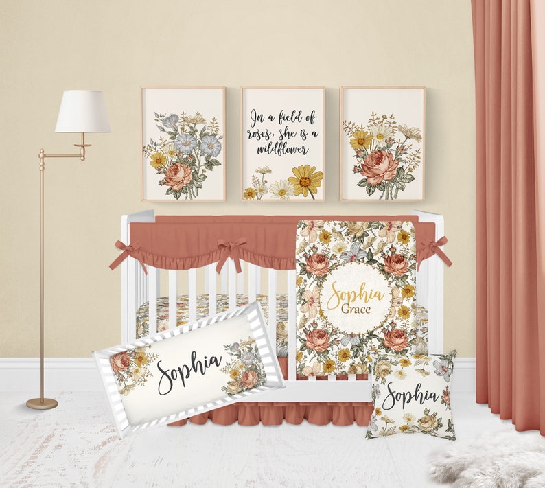 Op de afbeelding: Een wit babybedje met een bloemenbeddengoedset. De beddengoedset bevat een deken, een kussen en een bumper. Het babybedje is versierd met drie ingelijste bloemenprints. De prints tonen een bloemenpatroon met de tekst "In een veld vol rozen is ze een wilde bloem". Het babybedje is ook versierd met een kussen met de tekst "Sophia Grace" en een kussen met de tekst "Sophia".