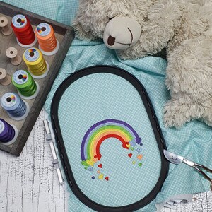 May include: A rainbow embroidery design with hearts on a light blue fabric in an embroidery hoop. The hoop is on a white wooden surface.