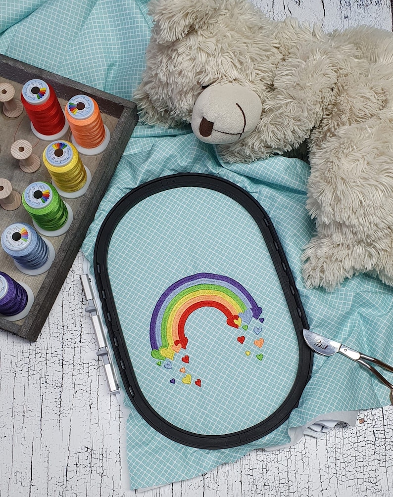 May include: A rainbow embroidery design with hearts on a light blue fabric in an embroidery hoop. The hoop is on a white wooden surface.