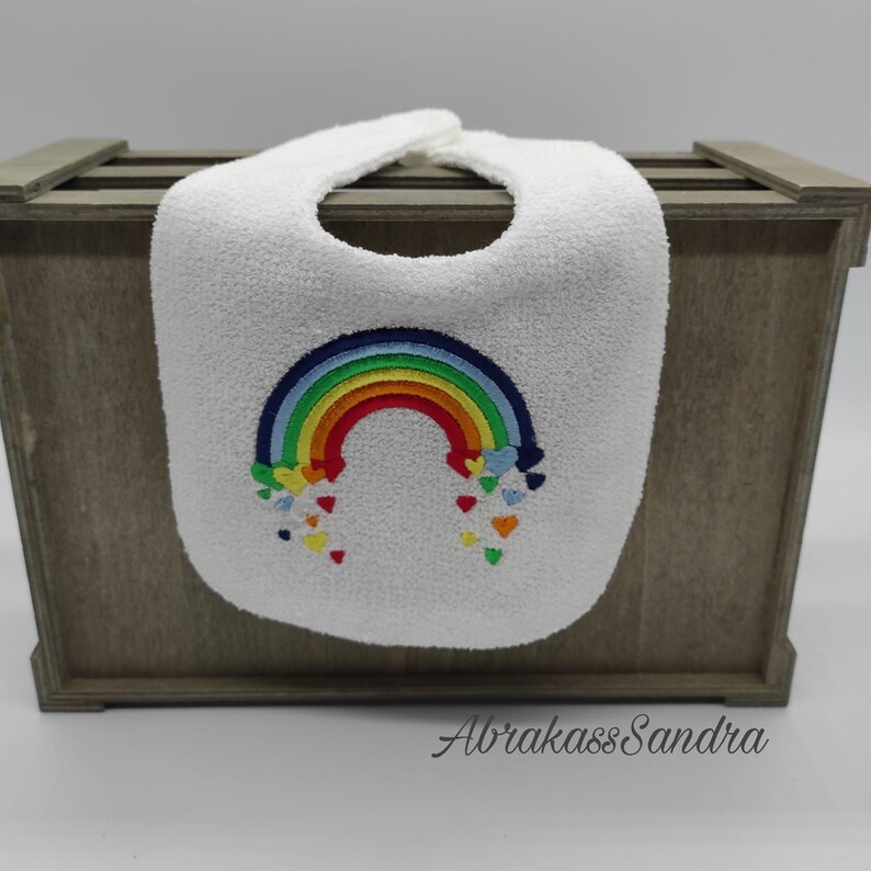 May include: White baby bib with a rainbow and hearts embroidered on it. The rainbow is made of red, orange, yellow, green, blue, and purple. The hearts are red, orange, yellow, green, blue, and purple.