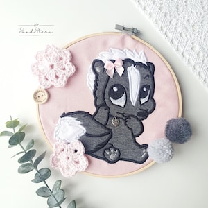 May include: Embroidered hoop art featuring a cute cartoon skunk with a pink bow, black and white fur, and a silver heart charm. The hoop is pink and surrounded by white crocheted flowers and gray pom-poms.