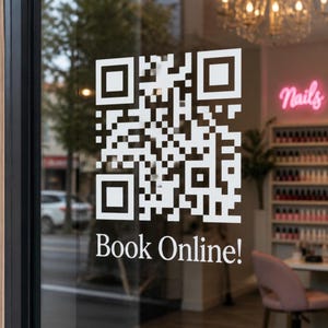 May include: A white vinyl decal applied to a glass window featuring a square QR code pattern and the text "Book Online!" in a serif font. The graphic is positioned on a transparent surface, allowing a view of a salon interior in the background.