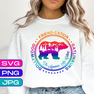 May include: White sweatshirt featuring a colorful graphic with a bear, mountains, and trees. The design includes the words "Friend-cation," "Dollywood," and "Gatlinburg." The phrase "The Mountains Are Calling" is also present.
