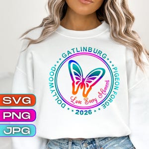 May include: White sweatshirt with a colorful butterfly design inside a circle. The design includes the words "Gatlinburg, Pigeon Forge, Dollywood" and "Love Every Moment 2026". The design is in shades of blue, green, orange, and purple.