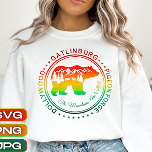 May include: White sweatshirt featuring a circular design with a bear silhouette and mountain scene in red, orange, and yellow. Text includes "Gatlinburg", "Pigeon Forge", "Dollywood", and "The Mountains Are Calling".