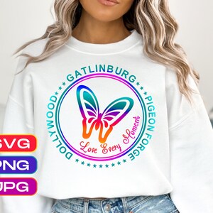 May include: A white crewneck sweatshirt featuring a circular graphic with a gradient butterfly in the center. The text reads Gatlinburg, Pigeon Forge, Dollywood, and Love Every Moment. The design uses a vibrant rainbow color gradient throughout.