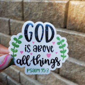 May include: A white sticker with the words "GOD is above all things" in black script, surrounded by green leaves and pink hearts. The text "PSALM 95:3" is at the bottom. The sticker has a holographic, glittery effect.