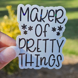 May include: A white sticker with the text "MAKER OF PRETTY THINGS" in black, with star accents. The sticker has a holographic, iridescent effect, reflecting various colors. The sticker is held up against a blurred green background.
