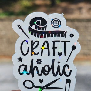 May include: A white sticker with the words "CRAFT aholic" in black script, surrounded by sewing-themed illustrations. The sticker features a tape measure, needle, scissors, and a safety pin. The design has a holographic, iridescent effect.
