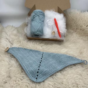May include: A light blue crochet project kit with a finished triangle scarf. The kit includes a ball of yarn, stuffing, a crochet hook, and a wooden button. The scarf is laid out on a fluffy, white surface.