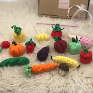 Op de afbeelding: Een verzameling gehaakte fruit en groenten, waaronder een ananas, een pompoen, een aardbei, een aubergine, een banaan, een wortel, een paddenstoel, een kers, een citroen, een appel, een ijshoorn en een paprika. De gehaakte items zijn gerangschikt op een wit, pluche oppervlak.