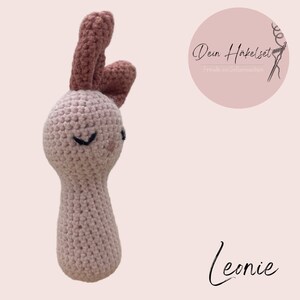 Puede incluir: Un sonajero de conejo de crochet rosa con orejas marrones. El conejo tiene una cara sonriente con ojos negros.