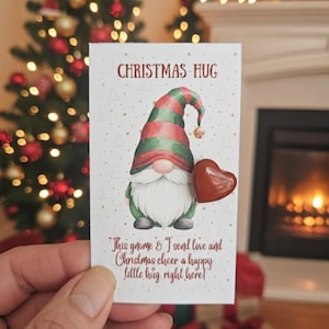 May include: A Christmas-themed card featuring a gnome holding a heart. The gnome wears a red, green, and white hat and has a white beard. The card reads "CHRISTMAS HUG" and a message about sending love and Christmas cheer.