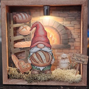May include: A wooden framed diorama featuring a gnome holding a loaf of bread. The scene includes a brick oven, various bread types, and a sign that reads "Victoria's Bakery". The diorama is illuminated with a small light.