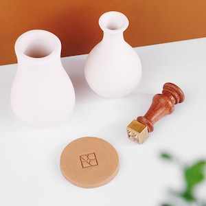 Peut inclure: Deux petits vases en céramique blanche avec des corps arrondis et des ouvertures étroites. Un tampon à cacheter en bois et en laiton et un dessous de verre rond en papier beige avec un motif géométrique sont également présents. Les articles sont disposés sur une surface blanche avec un fond marron.