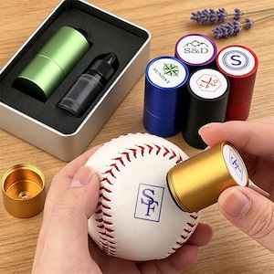 Op de afbeelding: Een witte honkbal met rode stiksels wordt gestempeld met een gouden cilindervormig hulpmiddel met de letters SF. Daarnaast liggen diverse andere cilindervormige stempels in groen, blauw, zwart, rood en paars, en een flesje zwarte inkt.