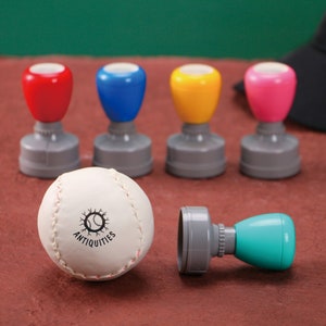 May include: A baseball and four colorful stamps with red, blue, yellow, and pink handles. The baseball is white with red stitching and the word "ANTIQUITIES" printed on it. The stamps have gray bases.