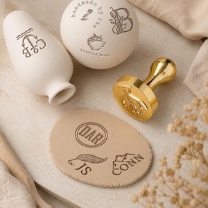 Può includere: Un vaso in ceramica bianca con ancora e incisione C&B, una sfera in ceramica bianca con testo handmade by rae, B e Bodega Mao, un timbro sigillo in metallo dorato con design di bicicletta e un disco di argilla beige con incisioni DAR, JS e CONN.