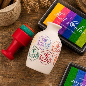 Könnte beinhalten: Eine weiße Keramikvase mit mehreren gestempelten sechseckigen Motiven, die Bienen und den Schriftzug "IZZY" zeigen. Ein roter Stempel mit grünem Schwamm und ein rechteckiges "Let's Color" Pigment-Stempelkissen liegen daneben.