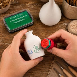 Könnte beinhalten: Eine weiße Keramikvase wird mit einem roten, sechseckigen Gummistempel mit Bienenmotiv und dem Text 1224 bestempelt. Die Vase zeigt grüne und blaue Bienenmotive mit dem Text 1224. Ein grünes Stempelkissen mit der Aufschrift Color Arts liegt daneben.
