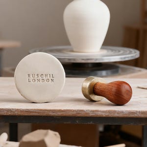 May include: A round, off-white pottery stamp with the text "RUSCHIL LONDON" is next to a wooden and brass tool. A white ceramic vase sits on a pottery wheel in the background. The items are on a wooden surface.