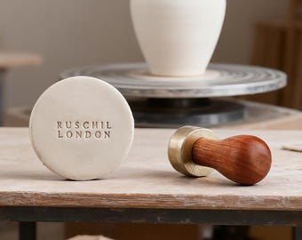 Puede incluir: Un disco de arcilla de color blanco roto con el texto RUSCHIL LONDON en relieve y un sello de latón con cabeza de metal pulido y mango de madera liso y redondeado, colocados uno al lado del otro sobre una superficie plana en un taller de cerámica.