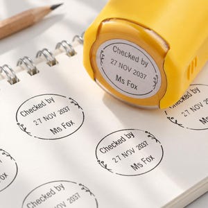 Op de afbeelding: Een gele zelfinktende stempel met een ronde voet, die zwarte inkt-afdrukken op papier achterlaat. De afdruk bevat de tekst "Checked by 27 NOV 2037 Ms Fox" omringd door een bloemmotief. De stempel ligt op een notitieblok met spiraalbinding.
