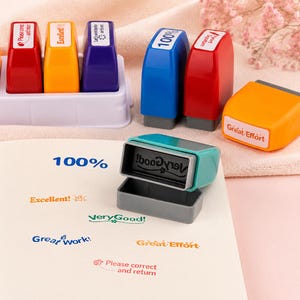 May include: A collection of colorful plastic self-inking stamps in red, yellow, purple, blue, and teal. The stamps feature various phrases including 100%, Excellent!, Very Good!, Great Work!, Great Effort, and Please correct and return.