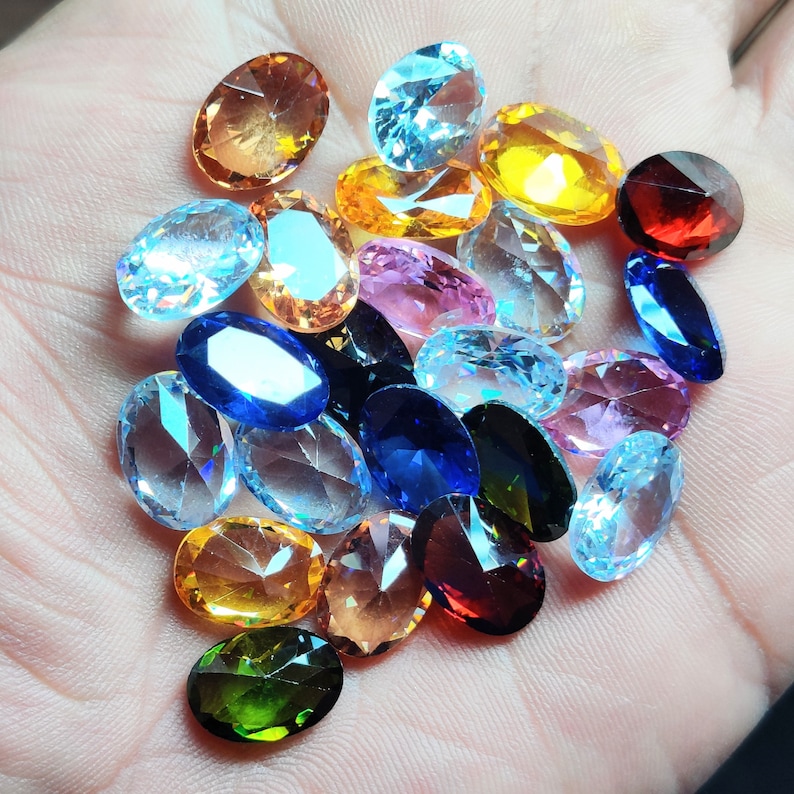 Op de afbeelding: Een verzameling ovaal geslepen edelstenen in diverse kleuren, waaronder oranje, geel, blauw, roze en groen. De edelstenen worden in een hand gehouden, waarbij hun facetten en glans worden getoond. Geschikt voor het maken van sieraden.