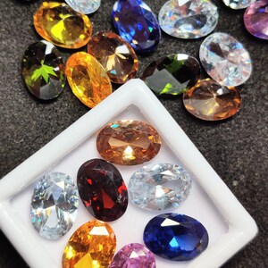 Op de afbeelding: Een assortiment ovaal geslepen edelstenen in diverse kleuren, waaronder geel, oranje, blauw en helder. Sommige edelstenen worden gepresenteerd in een witte plastic tray. De edelstenen zijn gefacetteerd en reflecteren het licht.