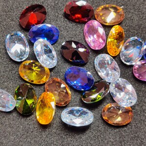 Op de afbeelding: Een assortiment ovaal geslepen edelstenen in diverse kleuren, waaronder rood, blauw, roze, geel en helder. De edelstenen liggen op een donkergrijze ondergrond, waardoor hun facetten en glans opvallen. Ideaal voor het maken van sieraden of knutselen.