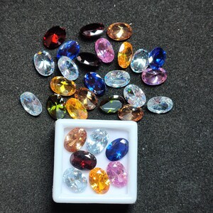Op de afbeelding: Een assortiment ovaal geslepen edelstenen in diverse kleuren, waaronder rood, blauw, geel en helder. Sommige edelstenen liggen verspreid op een donkere ondergrond, andere liggen in een witte vierkante container. De edelstenen hebben een gepolijst, reflecterend oppervlak.