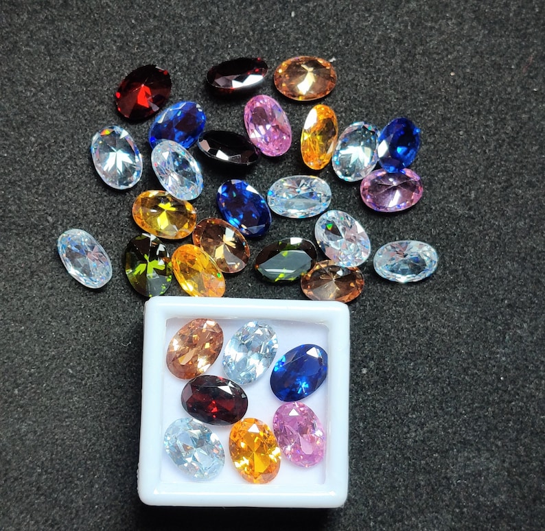 Op de afbeelding: Een assortiment ovaal geslepen edelstenen in diverse kleuren, waaronder rood, blauw, geel en helder. Sommige edelstenen liggen verspreid op een donkere ondergrond, andere liggen in een witte vierkante container. De edelstenen hebben een gepolijst, reflecterend oppervlak.