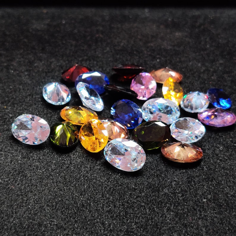 Op de afbeelding: Een verzameling ovaal geslepen edelstenen in verschillende kleuren, waaronder blauw, roze, geel en groen. De edelstenen zijn verspreid op een donkere ondergrond, waardoor hun facetten en glans worden benadrukt. De edelstenen zijn van verschillende grootte.