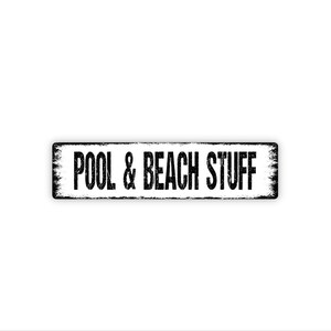May include: A rectangular black and white sign with the words "POOL & BEACH STUFF" in bold, distressed font. The sign has a weathered, vintage appearance, suggesting outdoor use. The sign is designed for pool and beach decor.