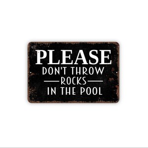 May include: A rectangular black metal sign with a distressed, rusted edge finish. The sign features white text that reads: PLEASE DON'T THROW ROCKS IN THE POOL. The lettering is centered, with the word PLEASE in a larger font size at the top.