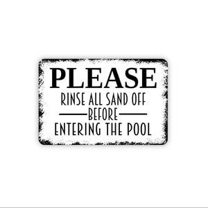May include: A rectangular white sign with a distressed black border and black text that reads: PLEASE RINSE ALL SAND OFF BEFORE ENTERING THE POOL. The sign features a clean, bold font for the word PLEASE and a thinner font for the remaining text.