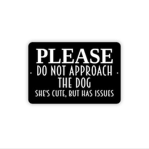 May include: A rectangular black sign with white text that reads: PLEASE DO NOT APPROACH THE DOG. SHE'S CUTE, BUT HAS ISSUES. The sign features a clean, bold font for the primary message and a thinner, sans-serif font for the secondary text.