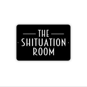 May include: A rectangular black sign with rounded corners featuring white text that reads THE SHITUATION ROOM in a clean, sans-serif font. The text is centered with a horizontal line above the word THE and small dots on the left and right sides.